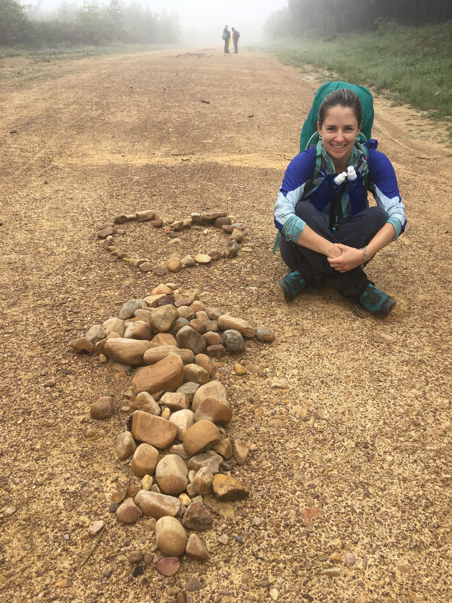 Woman sitting down next to a pile of stones shaped like an arrow pointing to a love heart whilst on the Camino De Santiago